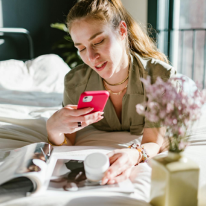 Woman lying in bed using her smartphone with flowers in the foreground and magazine nearby.